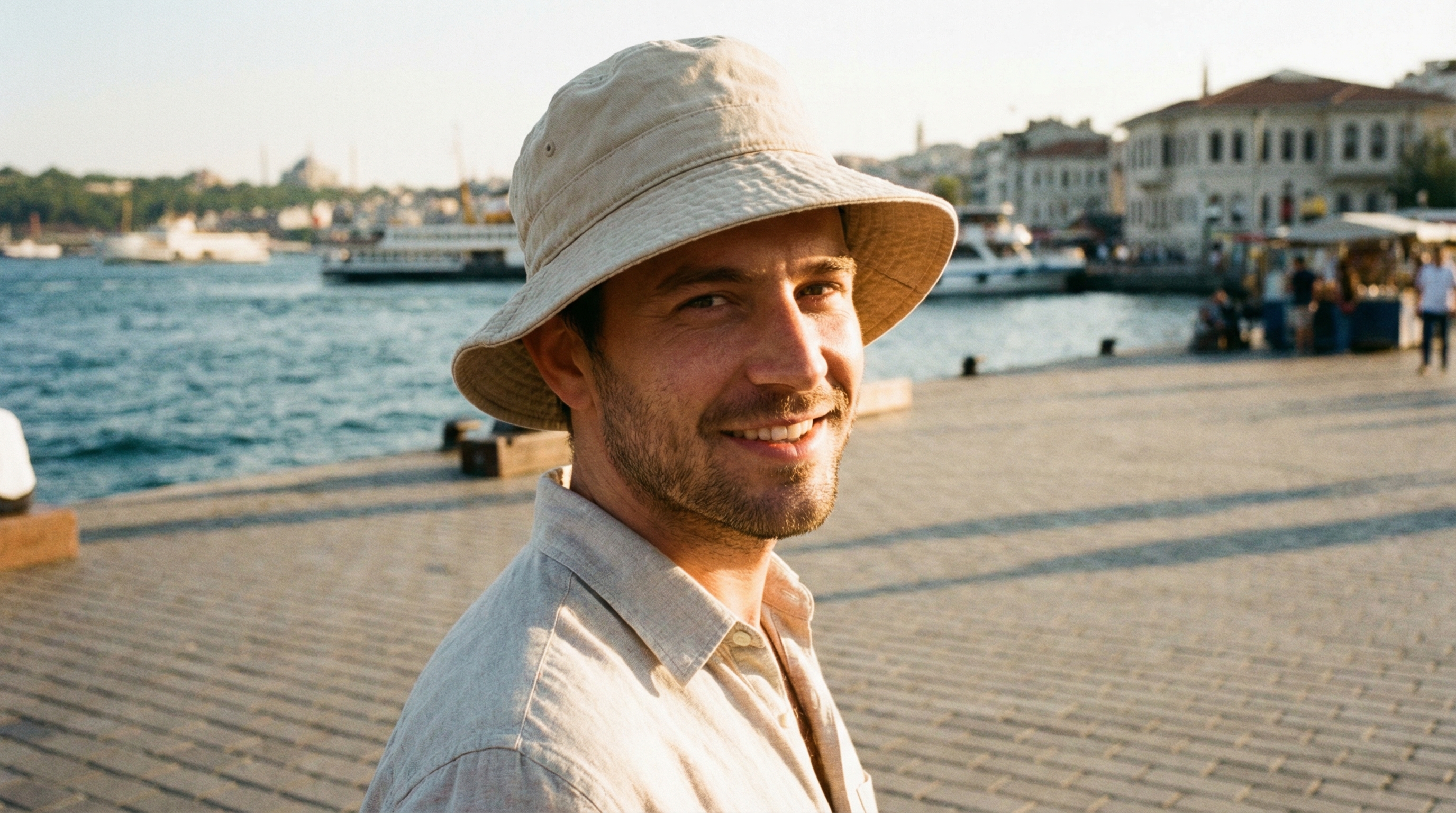 Patient portant un chapeau de pêcheur ample en coton clair après une greffe de cheveux, en extérieur sous le soleil méditerranéen d'Istanbul avec le Bosphore en arrière-plan — le chapeau ne touche pas le sommet du crâne et offre une protection solaire complète du cuir chevelu greffé